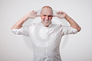 Mature european man closing his ears because of loud sound and noise. I do not want listen to you.