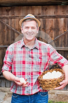 Mature farmer wearing hat while carrying fresh eggs in basket in barn