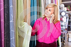 Mature female choosing draperies, holding smartphone in the store