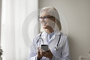 Mature female doctor standing at workplace with smartphone