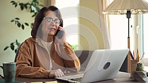 Mature female employee talk to client about order service in phone call, check laptop screen. Senior