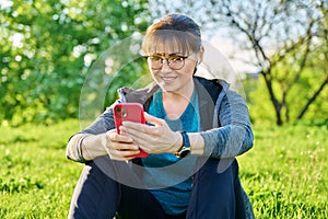 Mature female in headphones with smartphone watching video, sitting on grass