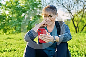 Mature female in headphones with smartphone watching video, sitting on grass