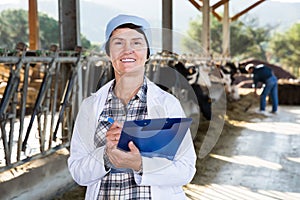 Mature female milker with clipboard standing at cowshed