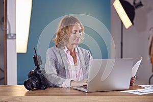 Mature Female Photographer At Desk With Laptop Checking Brief Before Fashion Shoot In Studio