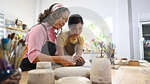 Mature female potter teaching young woman creating and sculpting clay pot in craft studio workshop