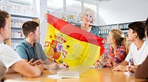 Mature female teacher shows the flag of Spain to teenagers and talks about this country during lesson at library school