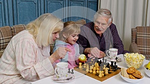 Mature grandmother grandfather with child girl grandchild playing chess game with on table in room