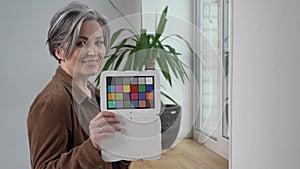 Mature gray haired businesswoman holds color checker smiling at camera. Woman in brown jacket shows target for color