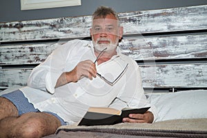 Mature, gray-haired man with beard and eyeglasses in hand, in pajamas, lying in bed looking at the camera while reading a book