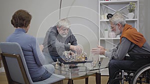 Mature grey-haired Caucasian men playing chess and talking as woman knitting in armchair. Elderly ill and disabled