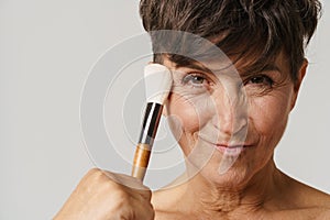 Mature half-naked woman smiling while using powder brush