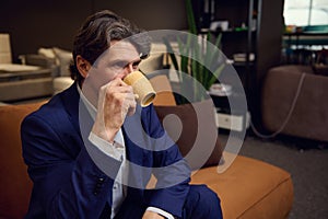 Mature handsome Caucasian man drinking coffee, sitting on a sofa in the furniture store showroom. Interior design, house