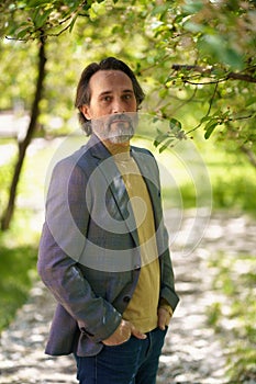Mature handsome man with grey beard looking thoughtful at the camera wearing casual jacket and jeans holding hands in
