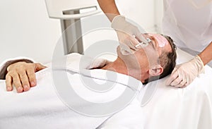 Mature handsome man lying down on a massage table at male beauty treatment for anti-aging and rejuvenation. Beautician applying