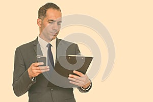 Mature handsome Persian businessman reading clipboard while holding mobile phone