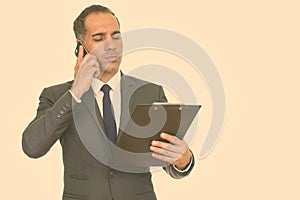 Mature handsome Persian businessman reading clipboard while talking on mobile phone