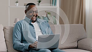 Mature happy african american man sitting on sofa at home relaxing smiling chatting on laptop via webcam talking on