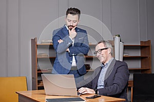 Mature happy businessman with grey hair in dark blue suit with his partner are thinking by the laptop in the office