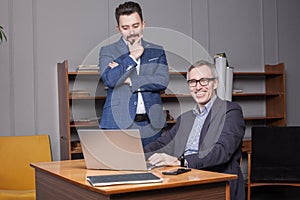 Mature happy smiling businessman in dark blue suit with his partner are standing by the laptop in the office and thinking about