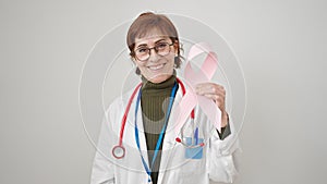 Mature hispanic woman doctor holding breast cancer awareness pink ribbon over isolated white background