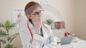 Mature hispanic woman doctor standing with doubt expression thinking at clinic