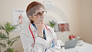 Mature hispanic woman doctor standing with doubt expression thinking at clinic
