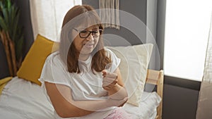 Mature hispanic woman holding her elbow in discomfort while sitting on a bed indoors with cozy bedroom decor photo