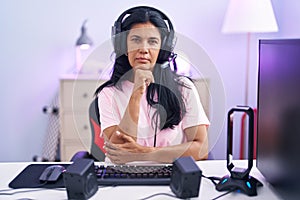 Mature hispanic woman playing video games at home looking confident at the camera with smile with crossed arms and hand raised on