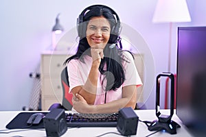 Mature hispanic woman playing video games at home looking confident at the camera smiling with crossed arms and hand raised on