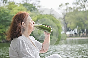 Mature Hispanic woman sitting in a park drinking green juice from a glass bottle