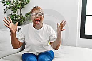 Mature hispanic woman sitting on the sofa at home crazy and mad shouting and yelling with aggressive expression and arms raised