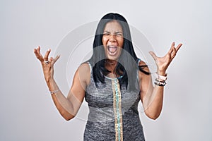 Mature hispanic woman standing over white background crazy and mad shouting and yelling with aggressive expression and arms raised