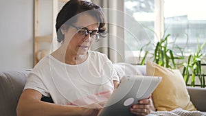 Mature housewife having time with tablet sitting on sofa in home interior.