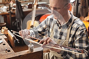 Mature instrument maker inside a rustic workshop is skillfully heating the hair of a violin bow to adjust its length. Blurred back