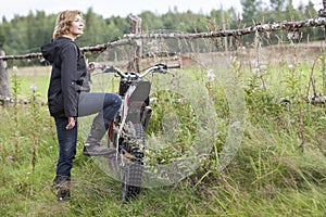 Mature joyful countrywoman standing with dirt motorcycle on rural road, copyspace