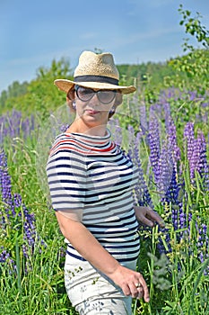 A mature lady in a hat and sunglasses stands among blooming lupins
