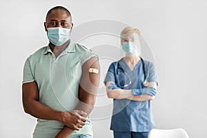 Mature lady nurse or doctor in uniform, protective mask stands with millennial african american patient