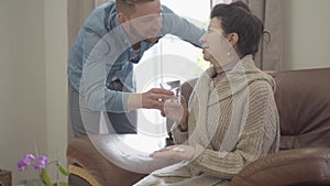 The mature lady sitting on the leather armchair at home, she is sick. Adult grandson brings the glass of water and gives