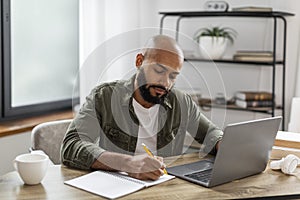 Mature latin man looking for job online, sitting at table, using laptop and taking notes, home interior, copy space