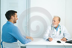 Mature male adult doctor interviewing smiling male patient and writing prescription sitting at table in medical office.