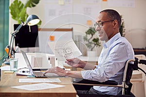 Mature Male Architect In Wheelchair Working In Office At Desk Studying Plans For New Building