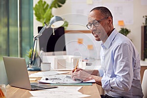 Mature Male Architect Working In Office With Model On Desk Studying Plans For New Building