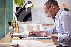 Mature Male Architect Working In Office With Model On Desk Studying Plans For New Building