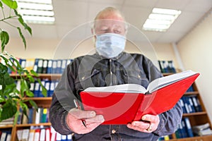 Mature male archivist holding open notebook in hands, looking at camera, man wearing face mask due Covid-19 pandemic. Focus is on