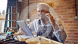 Mature Male Carpenter In Garage Workshop With Digital Tablet Talking On Mobile Phone
