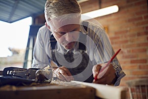 Mature Male Carpenter In Garage Workshop Marking Wood With Pencil