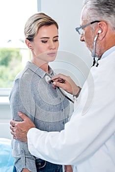 mature male doctor examining female patient by stethoscope