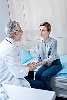 mature male doctor holding hands of female patient in hospital