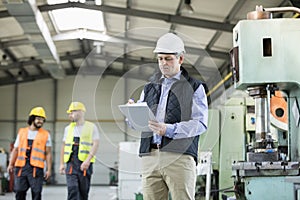 Mature male inspector writing on clipboard while workers in background at industry